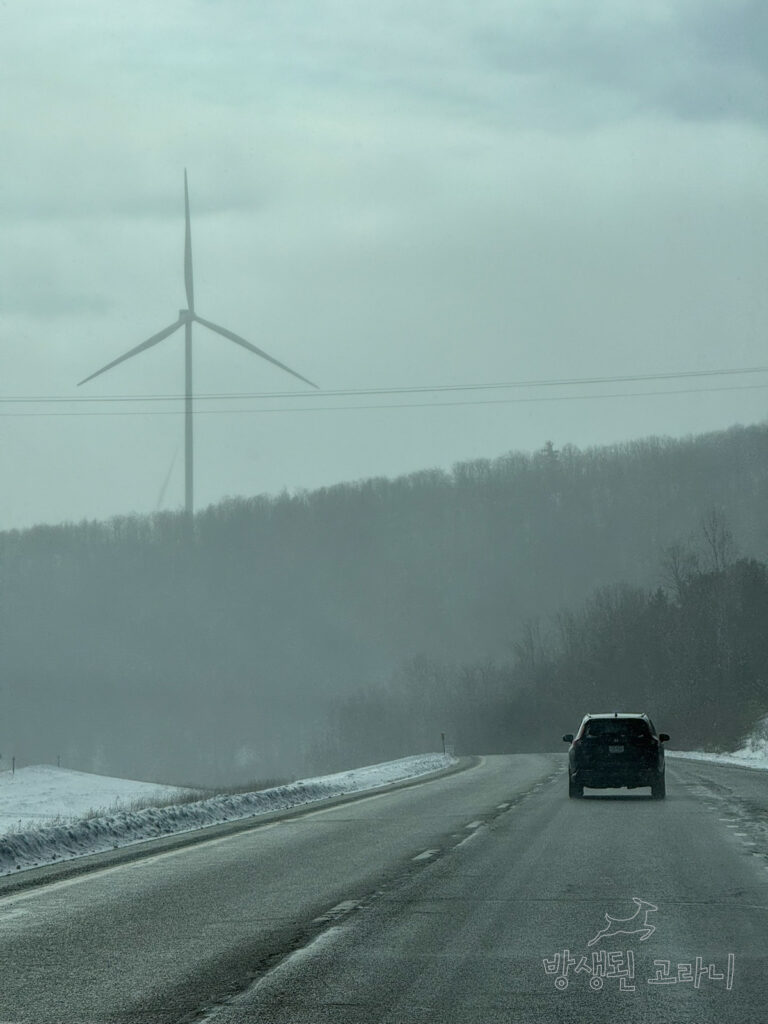 도로를 달리는 차 너머로 보이는 눈 덮인 산과 풍력발전기